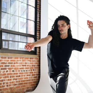 In a sunlit Somatic Lab with large windows and brick walls, Kiefer Josephs, with long curly hair, dances gracefully. Clad in a black shirt and pants, they extend one arm while bending the other above their head in a harmonious flow.