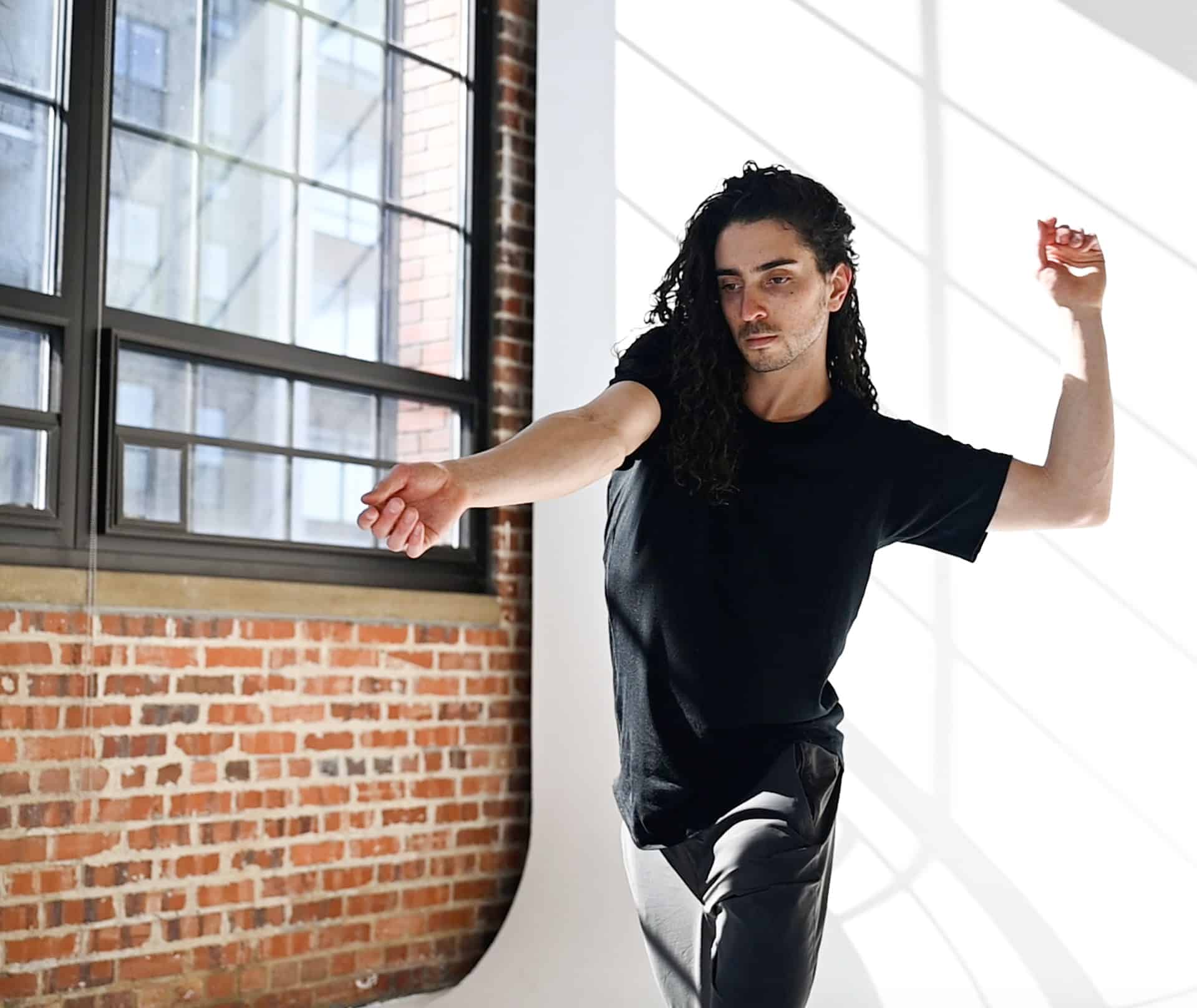 In a sunlit Somatic Lab with large windows and brick walls, Kiefer Josephs, with long curly hair, dances gracefully. Clad in a black shirt and pants, they extend one arm while bending the other above their head in a harmonious flow.