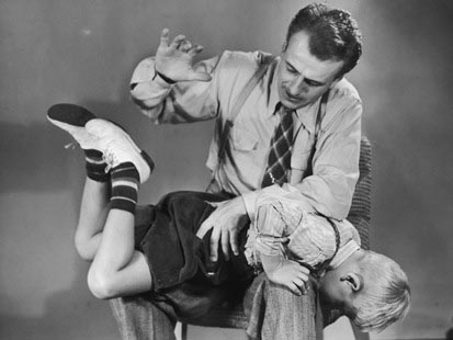 A black and white photo captures a father in vintage attire sitting on a chair, waiting to discipline his young son. The boy is bent over his father's knee, head down, arms hanging, embodying a timeless moment of home discipline.
