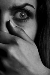 Black and white image of a person's face partially obscured by their hand. One wide eye is visible, with a solemn expression. The lighting creates high contrast, emphasizing the intensity of the gaze and the expressive details of the skin, as if a story is written across it.