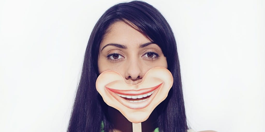 A woman with long dark hair holds a cardboard cutout of an exaggerated smiling mouth in front of her face, hinting at the effects of a fake smile. The background is plain white.