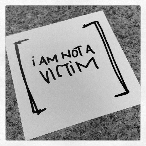 A note on a textured surface reads "[I am not a victim]" in bold black handwritten letters, embodying empowerment and personal growth. The image is captured in striking black and white tones.