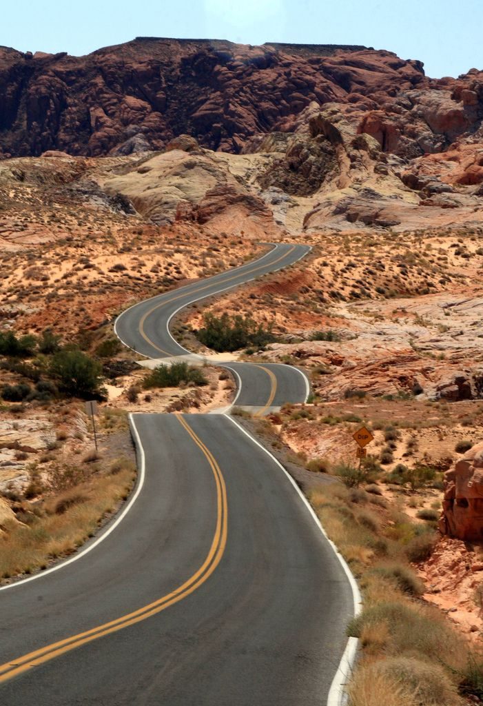 The winding road seems to drop out of sight as it stretches through a desert landscape, flanked by red rocky hills and dry vegetation under a vast blue sky. The yellow center lines guide travelers through this rugged terrain, inviting the adventurous to drop in and explore.