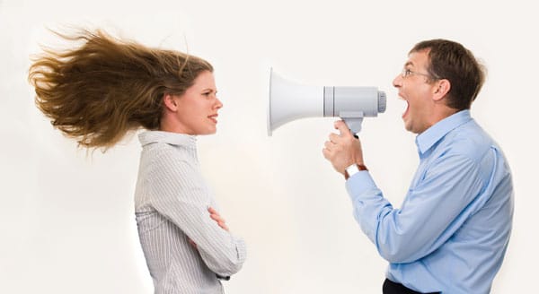 A man in a blue shirt holds a megaphone, shouting with a commanding tone of voice towards a woman with long hair blowing back. She stands defiantly with her arms crossed, wearing a striped shirt against the plain background.