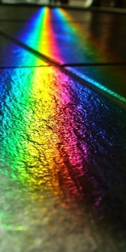 A vibrant rainbow, almost like a moment from a Kurt Wimberger painting, reflected on a textured, tiled floor. The colors transition smoothly from red through violet, creating a vivid display across several tiles.