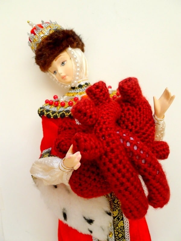 A doll dressed in ornate medieval-style clothing, featuring a crown and red robe, holds a red knitted heart organ symbolizing "Mend Your Mind." Against a plain white background, the intricate details of the attire and the knitted piece are highlighted, reflecting Vija Rogozina's thoughtful artistry.