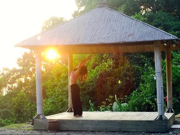 A person practices neurosculpting yoga under a thatched-roof gazebo at sunrise or sunset, surrounded by lush green trees. The sun is partially visible, casting a warm glow over the serene scene.