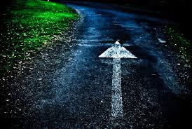A paved path with a white arrow pointing forward evokes Susan Aplin's notion of "Never Go Back". The left side is bordered by vividly green grass, while the right side lingers in shadow. This dimly lit scene creates a striking contrast between the bright grass and dark surroundings.
