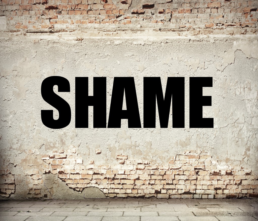The word "SHAME" is boldly painted in black on a weathered brick wall with a cracked, beige plaster surface. Crafted anonymously, the scene features a ground paved with gray bricks, adding to the urban, gritty atmosphere.