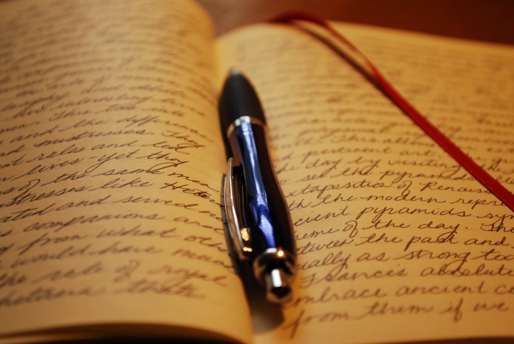 An open journal with handwritten cursive text and a black pen resting on the pages, reminiscent of notes from "Average" by Susan Aplin Pogue. A red ribbon bookmark adds charm to the warmly lit scene, creating a cozy and inviting atmosphere.