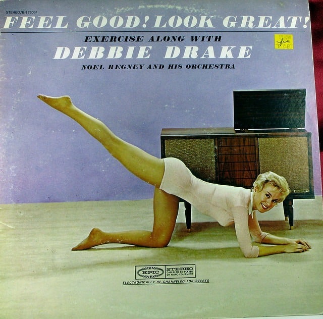 A woman in a pink leotard exercises on the floor, lifting one leg behind her, embodying 'Brain Happiness.' She smiles while stretching on a cream-colored carpet. In the background, a wooden console TV plays her favorite tracks from "Motivate Yourself to Exercise.