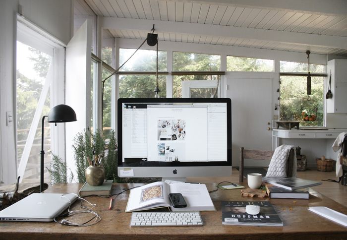 A cozy home office with a large wooden desk facing a window enhances workplace productivity. An iMac displaying a collage, books, plants, a lamp, and a cup are neatly arranged. Natural light streams in, and trees are visible outside. White walls and a high ceiling create an airy feel.