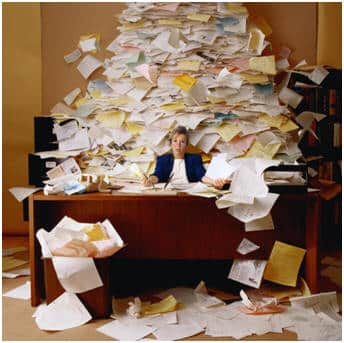A person sits at a cluttered desk, engulfed by an overwhelming tower of papers, creating a chaotic scene. Unnecessary organization seems evident as papers scatter across the floor and air, with a brimming trash bin beneath.