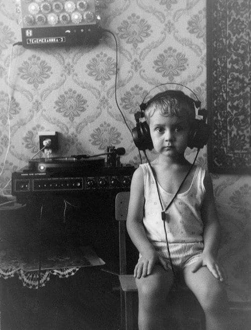 A young child, embodying the power of listening, sits on a chair in front of a record player and audio equipment. Dressed in a sleeveless top and shorts against vintage wallpaper, the child gazes directly at the camera through large headphones.
