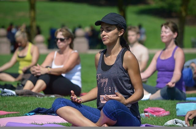 Michelle Lee Weldon, in a cap and tank top, sits cross-legged on a yoga mat, meditating peacefully in a park. Engaged in her yoga practice amidst others, she basks in the sunny day with grass and trees providing a tranquil backdrop.