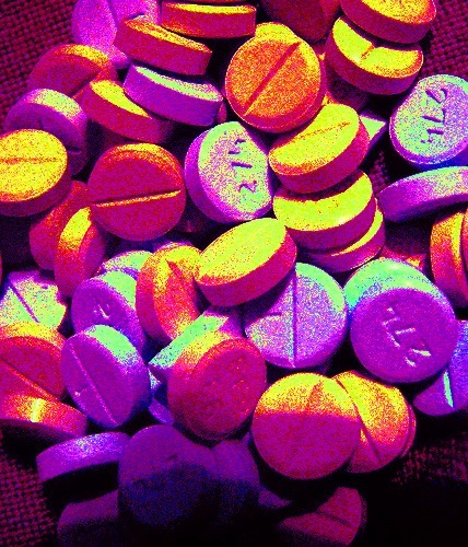 A pile of round pills with a psychedelic, colorful filter conjures a sense of meditation. The pills are mainly purple, orange, and yellow, with various lighting effects creating a vibrant, glowing appearance. Some pills have a line or symbol on one side.