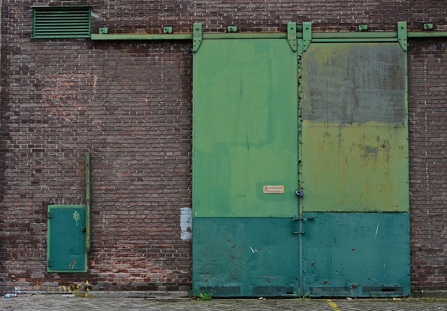 A large green metal sliding door on a brick building bears the marks of age and rust, reminiscent of NSI Warrior 1's resilient armor. To the left, a smaller utility box adds character beneath a vent near the top left. Cobblestones pave the ground, adding to this urban fortress ambiance.