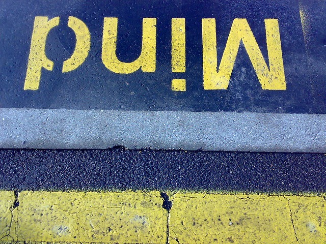 Yellow text reading "Mind" is painted upside down on asphalt, next to a concrete curb and a yellow line on a textured surface, possibly at a train station or platform edge, serving as a cautionary warning. This subtle reminder embodies the power of the mind to influence awareness and safety.