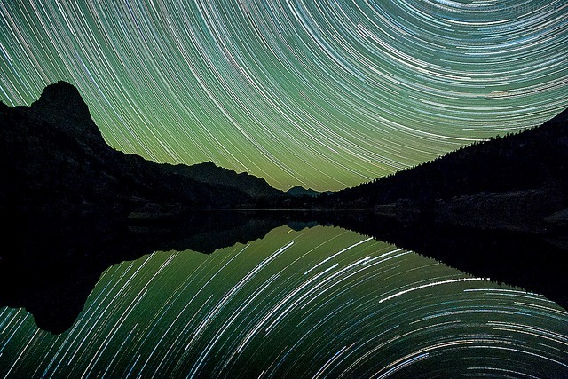 Renata Di Virgilio's mesmerizing long-exposure photograph captures circular star trails above a mountainous landscape. The trails reflect on the lake's calm surface, creating a symmetrical and serene visual effect that embodies the tranquility of a certified Facilitadora de Neurosculpting experience.