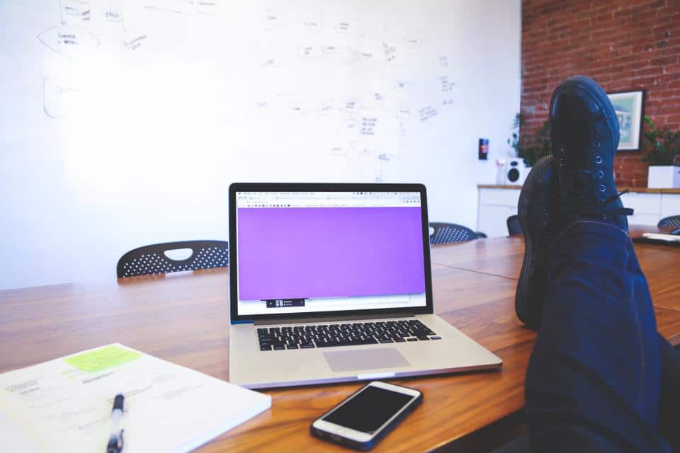 A person embraces workplace mindfulness with their feet up on a table in a conference room, gazing at a laptop with a purple screen. A smartphone and notepad sit nearby, while the background reveals a whiteboard with notes on a brick wall, suggesting an oasis of calm amidst the office buzz.