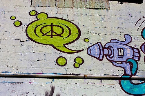 Graffiti on a brick wall depicts a cartoon-style futuristic gun shooting a green speech bubble with a purple peace symbol inside. The artwork is colorful and playful, capturing the spirit of emotional warriors spreading tranquility through bursts of vibrant creativity.
