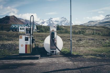 A rural scene showcases two vintage gas pumps under a vast blue sky, echoing the nostalgic cry of "Fill 'Er Up!" In the background, Travis Rumsey's humble building sits serenely against the grassy expanse and looming mountains.