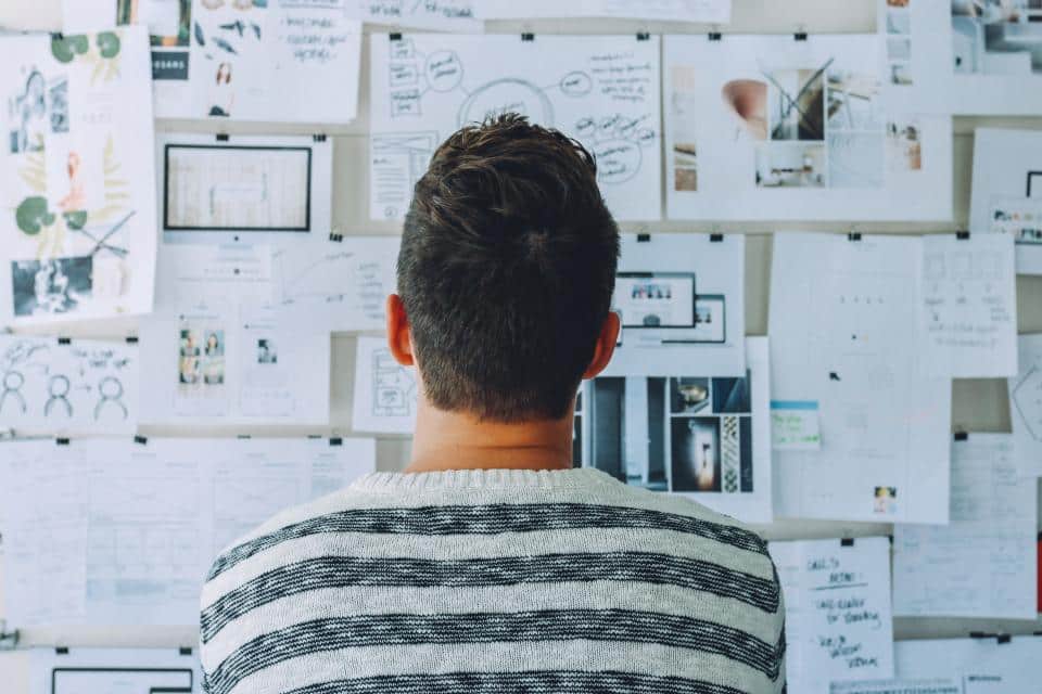 A person wearing a striped sweater looks at a wall covered with papers, sketches, and diagrams. The scene suggests brainstorming or planning, with various notes and images pinned up for reference.
