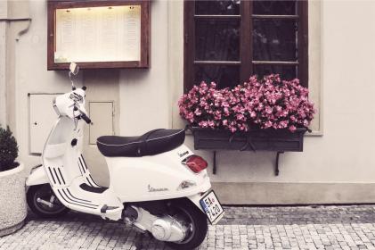 A white scooter, perfect for a vacation adventure, is parked on a cobblestone street beside a building with a wooden-framed menu on the wall. A window box filled with pink flowers sits under a nearby window, inviting you to enjoy every day’s simple pleasures.