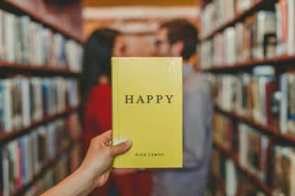 In a library aisle, someone clutches Megan Winkler's yellow book titled "HAPPY," while in the blurred background, two people stand close, engaging deeply between shelves full of books, perhaps delving into both meditation and relationship.