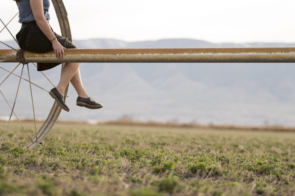 Cynthia Beard sits on a large, horizontal metal pipe with a wheel-like structure, overlooking a grassy field and distant mountains. Her casual shoes dangle as she embraces the super-awesome tranquility of this meditative setting.