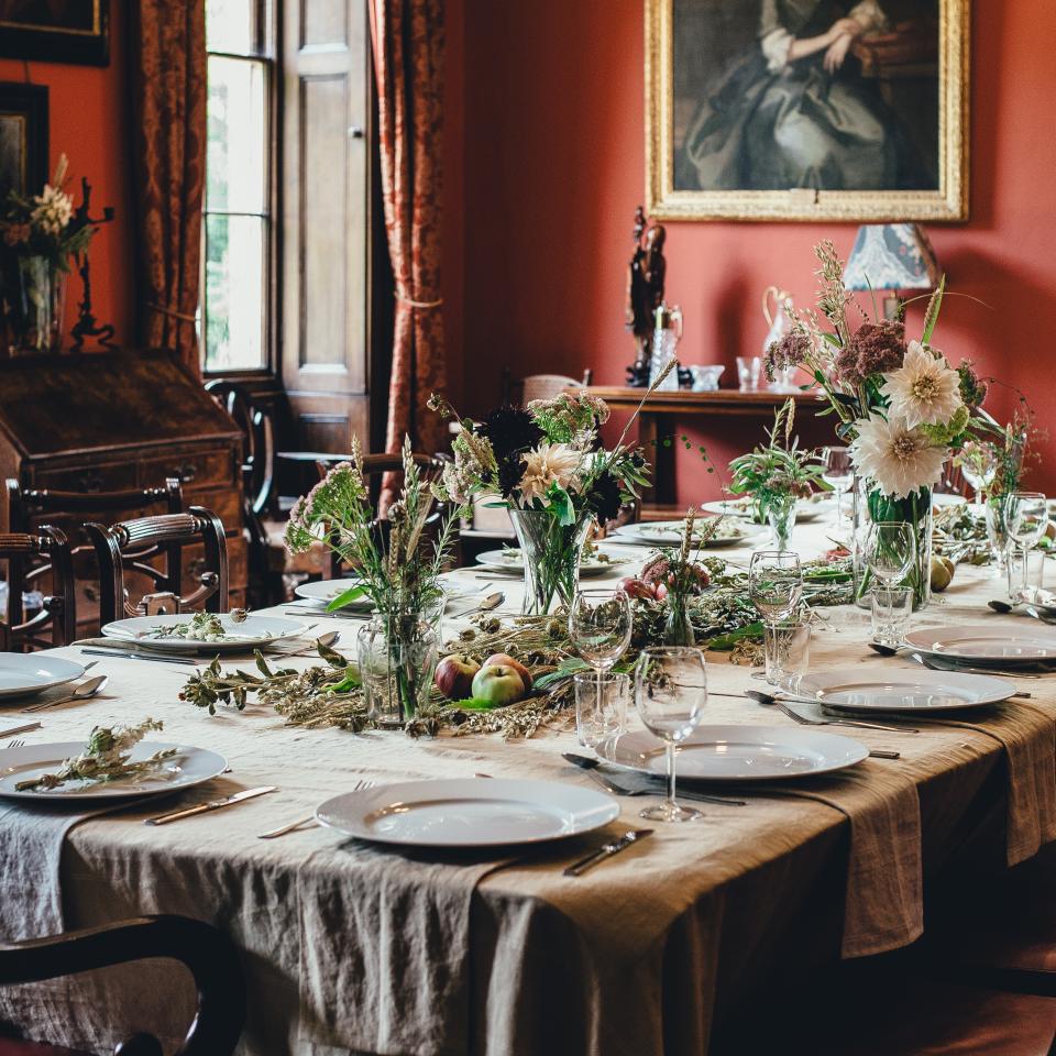 A dining table is elegantly set for a holiday meal with white plates, glassware, and a centerpiece of flowers and greenery. The room's red walls, vintage furniture, a portrait, and large window drapes evoke a classic, cozy atmosphere perfect for savoring festive moments.