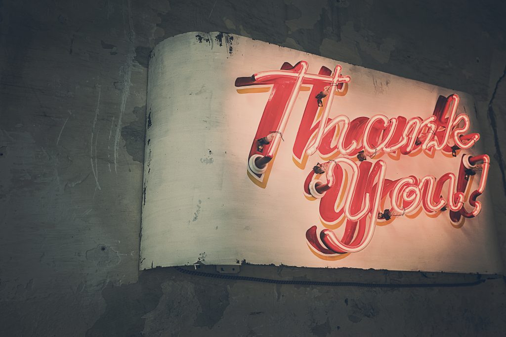 A vintage-style neon sign with the words "Thank You!" in red cursive letters is mounted on a textured, weathered wall, exuding a sense of gratitude. The background is dimly lit, adding an aged and retro feel to this nostalgic choice of decor.