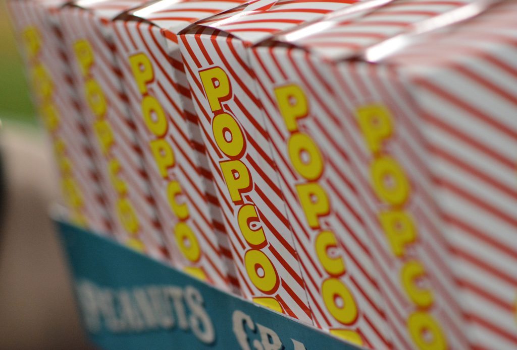Close-up of popcorn boxes with red diagonal stripes and yellow text saying "POPCORN," stacked in a row to create a pattern. It's like Lisa's Note of the Day tucked into memory, vivid yet elusive. Below, the edge of a blue box with partial text "PEANUTS" peeks through.