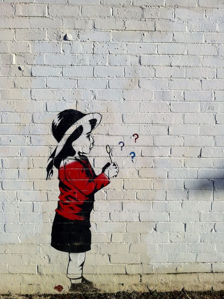 Street art on a white brick wall by Kelley Seriano depicts a young girl in a red coat blowing bubbles, which transform into red, blue, and purple question marks. The mural subtly whispers the message: Don't Be Afraid. Ask Questions.