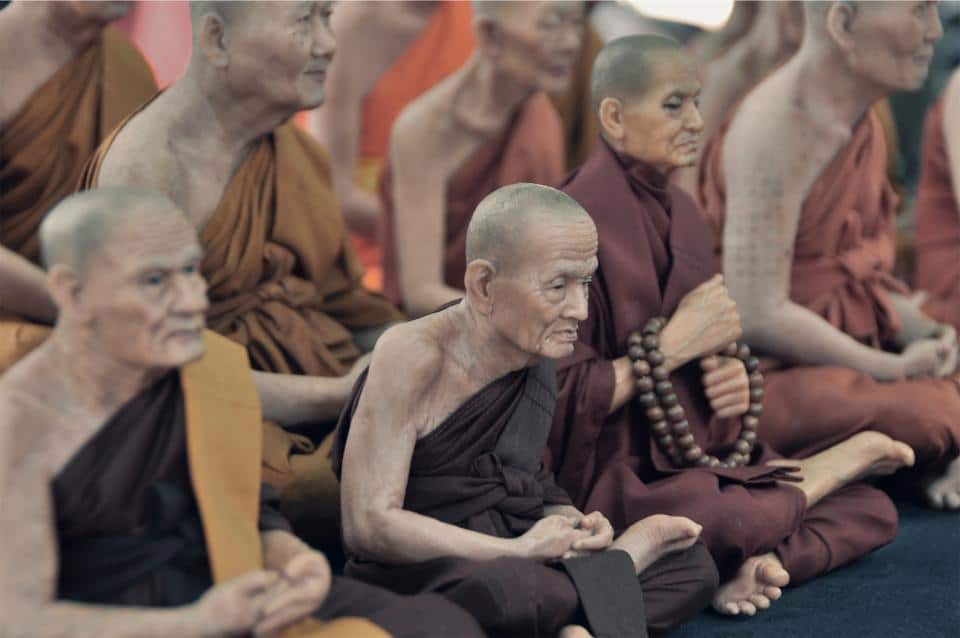 A group of elderly monks, embodying a "one size fits all" wisdom, sit cross-legged in traditional saffron and maroon robes. One monk, holding prayer beads, reflects the serene focus shared among them in their meditative posture.