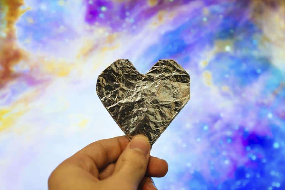 A hand holds a heart-shaped piece of crumpled foil, set against a colorful, abstract backdrop of swirling patterns in purple, blue, and pink—like an artistic message from Lisa's Note.
