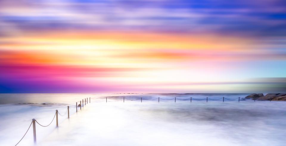 A dreamy, long-exposure shot of the ocean at sunset. Vibrant colors blend across the sky, reflecting on the water. A line of posts with a rope creates a pathway into the misty sea, as if each step takes you deeper into a ruled world of tranquility and surreal wonder.