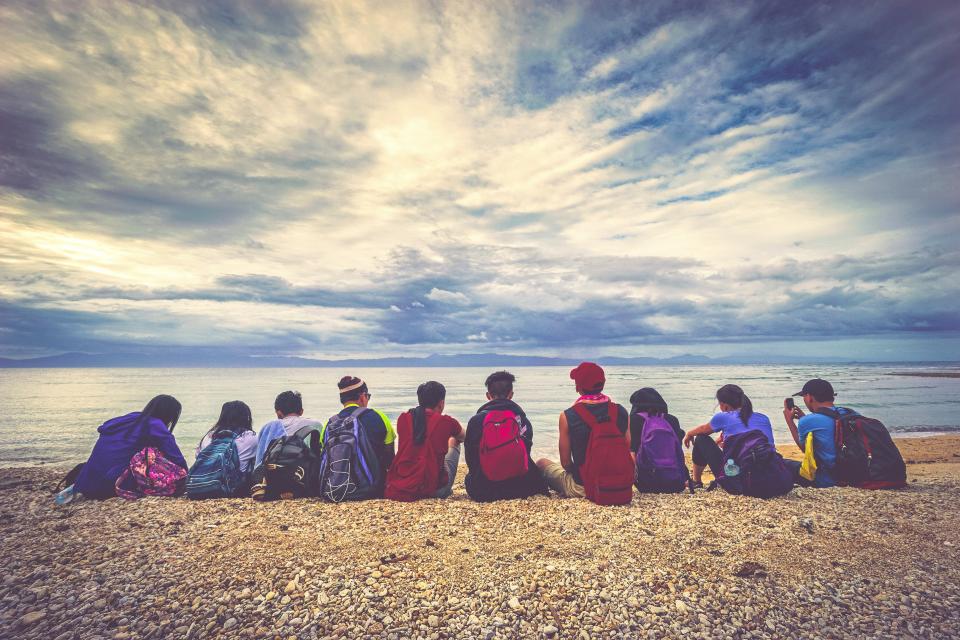 A group of nine people with backpacks sits on a rocky beach, facing the ocean. Part of an adventurous kids program, they enjoy the serene view under a sky filled with dramatic clouds and a distant horizon.