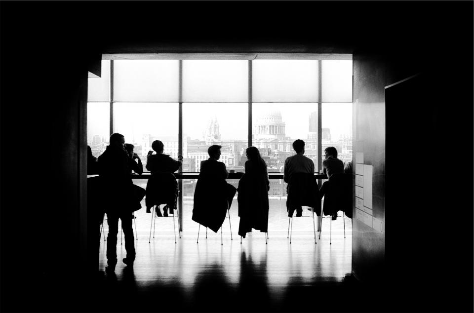 Silhouettes of seven people sitting and standing by a large window, overlooking a cityscape. The scene is in black and white, creating a contrast between the bright window and the dark interior.