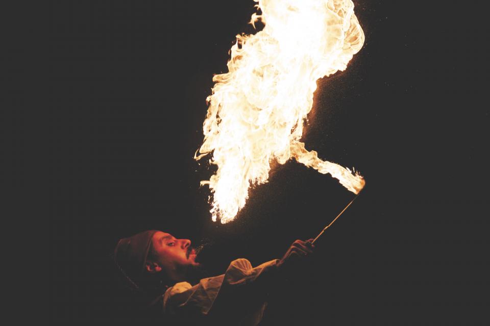 Cynthia Beard, a skilled performer, breathes a large plume of fire into the dark night sky, holding a lit torch. The flames create a dramatic and meditative effect against the black background as time seems to slow down.