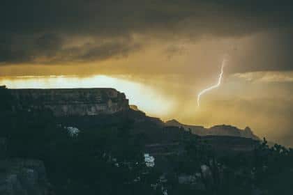 A dramatic landscape captured by Kelley Seriano shows a distant lightning bolt striking under a dark, stormy sky. The foreground features rocky terrain silhouetted against a yellowish glow from the sunset, expanding your experience of nature's raw beauty.