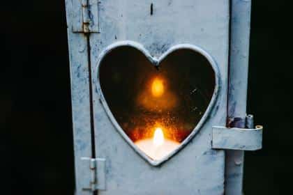 A white lantern with a heart-shaped window reveals a glowing candle inside against a dark background, like Lisa's Note guiding the way. The warm light of the flame contrasts with the cooler tones of the lantern's metal exterior.