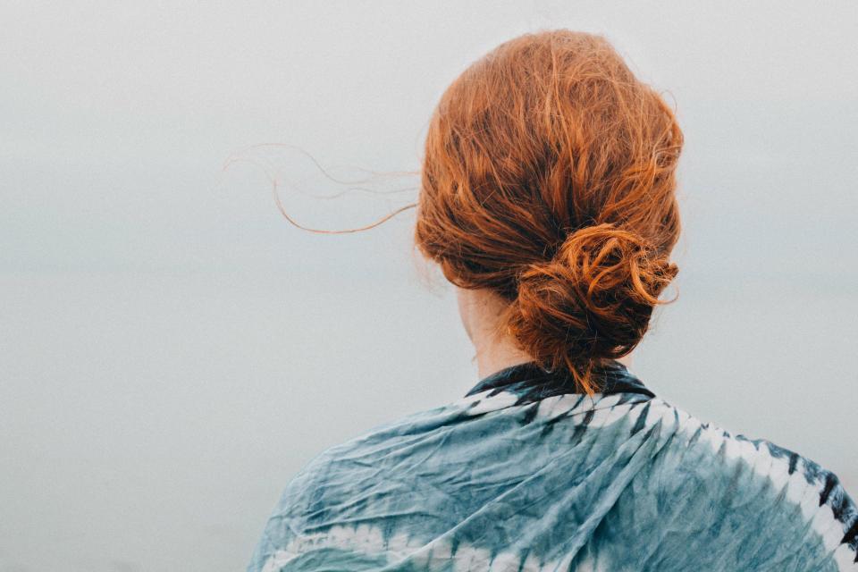 A mindful day unfolds as a person with red hair in a loose bun stands facing a misty landscape, wearing a blue and white tie-dye shawl. The air is foggy, their hair subtly blowing in the wind like Lisa's note to the earth.