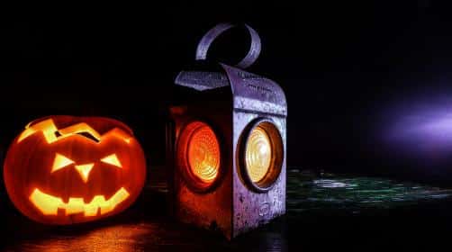 A glowing jack-o'-lantern with a carved face sits beside a rustic metal lantern on a dark surface. The scene, reminiscent of Travis Rumsey's novel, is illuminated by their orange lights, casting a warm glow against the black background.