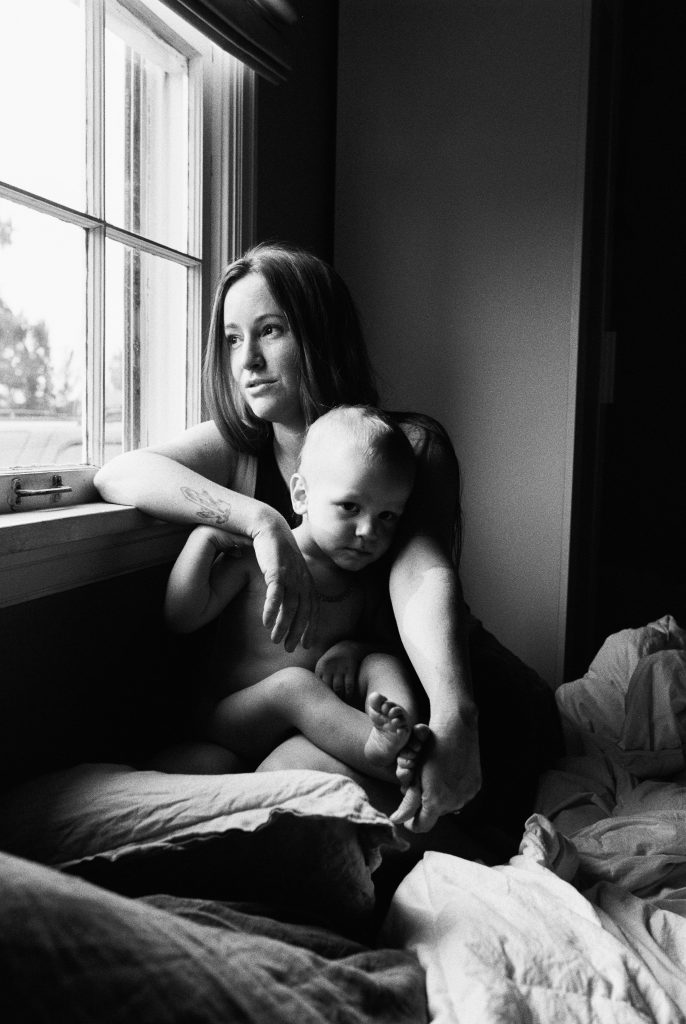 A woman with long hair sits beside a window, holding a baby on her lap. Both gaze outside with a serene expression, as if reflecting on the day's note. The black and white image captures this calm, intimate moment, with soft lighting highlighting their faces.