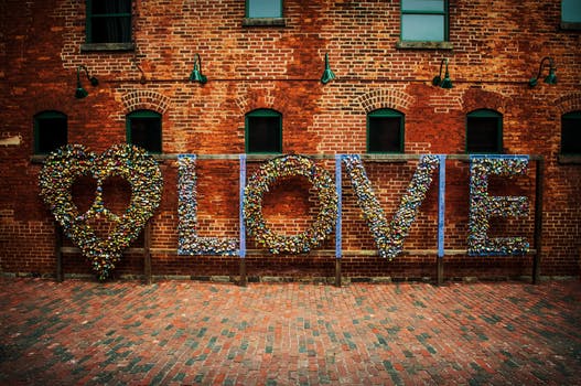 A large installation by Vija Rogozina against a red brick wall spells out "LOVE" with a heart shape. TRIGGERed by passion, the letters and heart are adorned with colorful locks, while the brick pavement below complements the rustic setting.