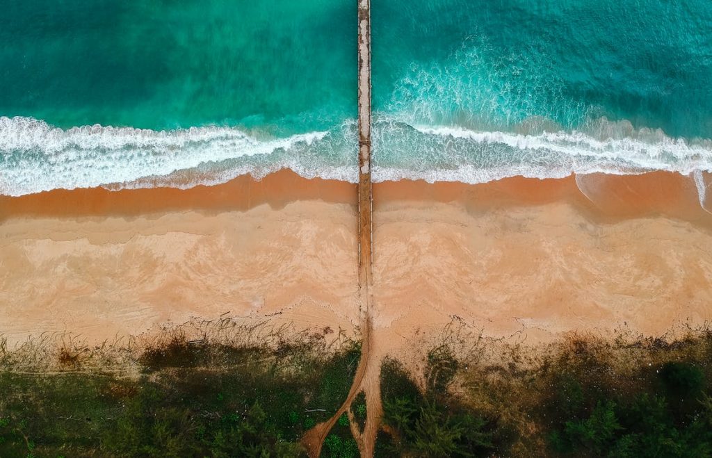 Aerial view of a beach divided by a straight narrow pier. Turquoise waves crash on the sandy shore, reminiscent of castles made of sand, while lush greenery lines the beach on the other.