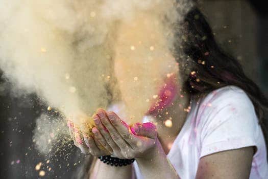 Jody Domerstad stands in a white shirt, blowing a handful of colorful powder, creating a vibrant cloud that echoes the joy of celebration. The scene embodies the spirit of "Talk to Your Dis-ease," emphasizing healing through color and festive expression.
