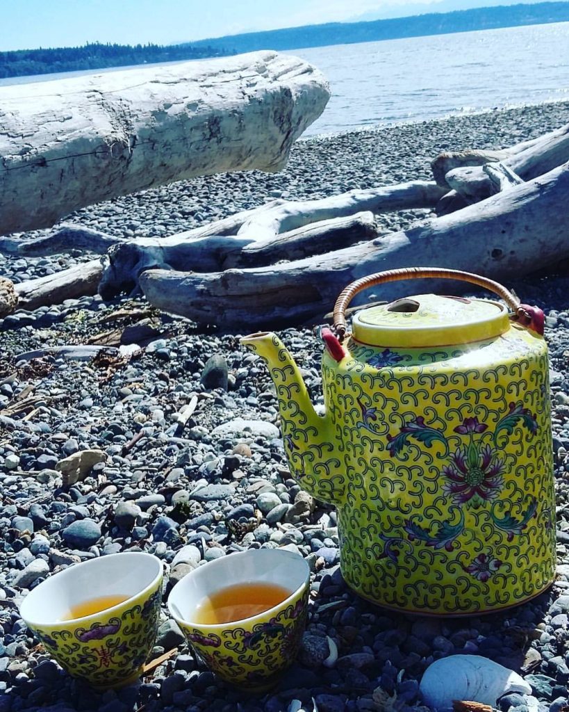A yellow teapot with floral designs and two matching cups filled with tea, reminiscent of Vija Rogozina's artistry, sit on a rocky beach. Driftwood is scattered nearby, while the sea and distant shoreline stretch under a clear blue sky.