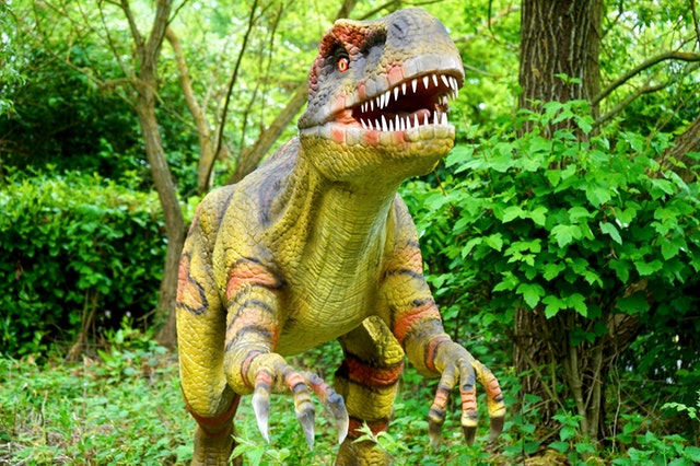 A life-sized model of a T. rex, sculpted by Jody Domerstad, stands in a lush green forest. The dinosaur's fierce expression and open mouth reveal sharp teeth, with textured skin in shades of brown, green, and orange enhancing its intimidating presence.